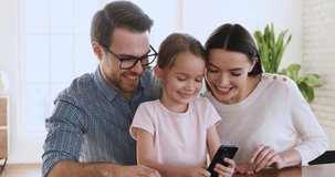 Cute small child daughter holding smartphone showing parents watch funny social media video take selfie together on modern phone using app, happy family having fun with technology sit at home table
