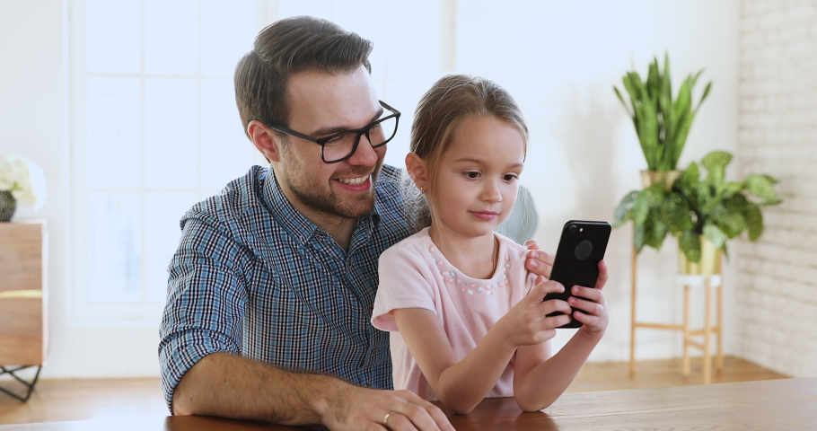 Young parent dad and cute small kid daughter bloggers recording lifestyle vlog on smartphone talking looking at phone using funny application having fun with tech education concept sit at home table Royalty-Free Stock Footage #1044455056