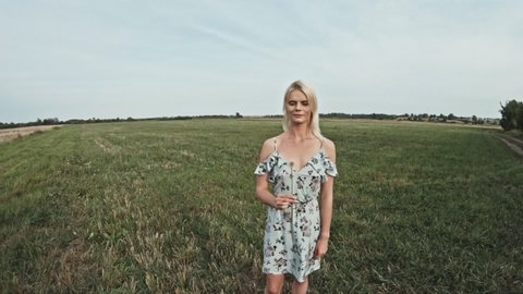 Portrait of blonde woman blowing a dandelion Flower wearing light casual clothes with landscape view of green field meadows and clear sky as background during the day