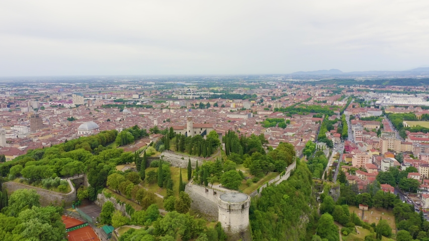 Brescia, Italy. Castello di Brescia, a medieval stone fortress with red-tiled rooftops and cylindrical towers, sits atop Cidneo Hill overlooking the city and surrounding mountain ranges. The aerial footage captures the fortified complex from a sweeping orbit, revealing its extensive stone walls, bastions, and the dense urban landscape below under an overcast summer sky. The castle, one of the largest fortified complexes in Italy, dominates the hillside with its imposing ramparts and historic architecture, offering a commanding view of the Lombardy plains and the distant Alps. The surrounding greenery and terracotta rooftops of the city create a striking contrast against the grey stone fortifications, Point of interest