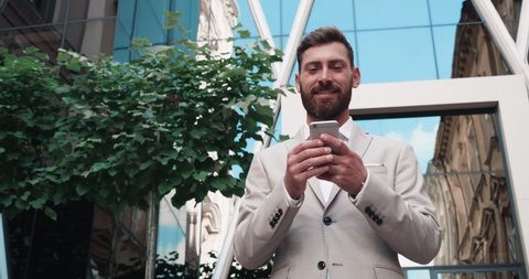  Businessman in a beige classical Suit browsing his Smartphone. Man  Walking by the High Glass Office Building and chatting on Social Media Video Stok