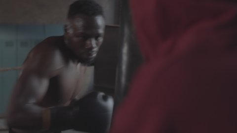 Waist-up shot of muscular Afro-American boxer practicing jabs and crosses on heavy punch bag at amateur boxing club and yelling while coach is holding bag and giving instructions