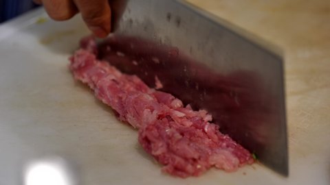 Close up chef's hand using cleaver knife chop minced raw pork on white plastic chopping block on the countertop in Asian restaurant's kitchen.