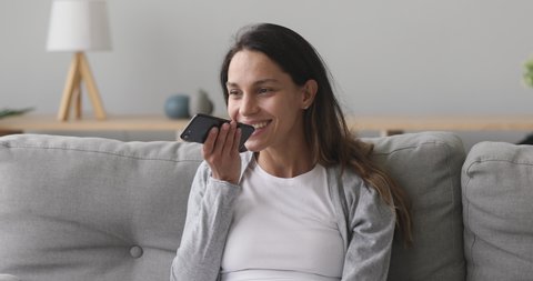 Happy millennial girl activating virtual assistant on mobile phone. Smiling young mixed race woman recording answering by audio message in social network for friend or using translator at home.