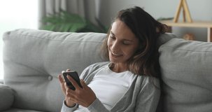 Happy young woman relaxing on cozy couch, using smartphone. Smiling millennial mixed race girl looking at cellphone screen, typing message for friend in social network, shopping online, web surfing.