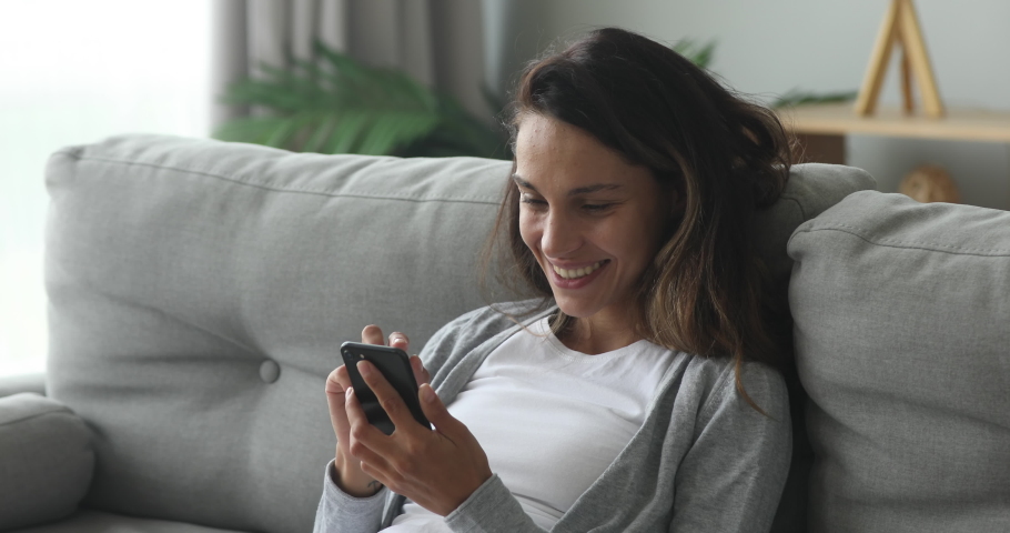 Happy young woman relaxing on cozy couch, using smartphone. Smiling millennial mixed race girl looking at cellphone screen, typing message for friend in social network, shopping online, web surfing. | Shutterstock HD Video #1044486907