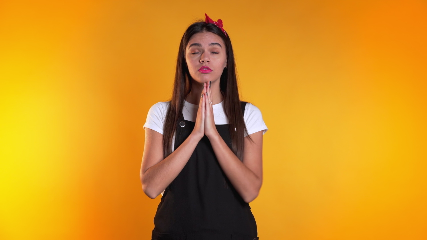 Cute european young girl in black overall praying over yellow background. Woman begging someone. 4k.