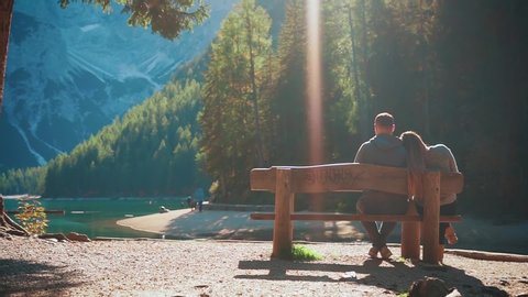 happy lover in wild nature, young couple sitting on wooden bench, two hike tourists relax travel, panoramic top hill rock view blue lake Braies Italy snow-capped mountains sun rays light, slow motion