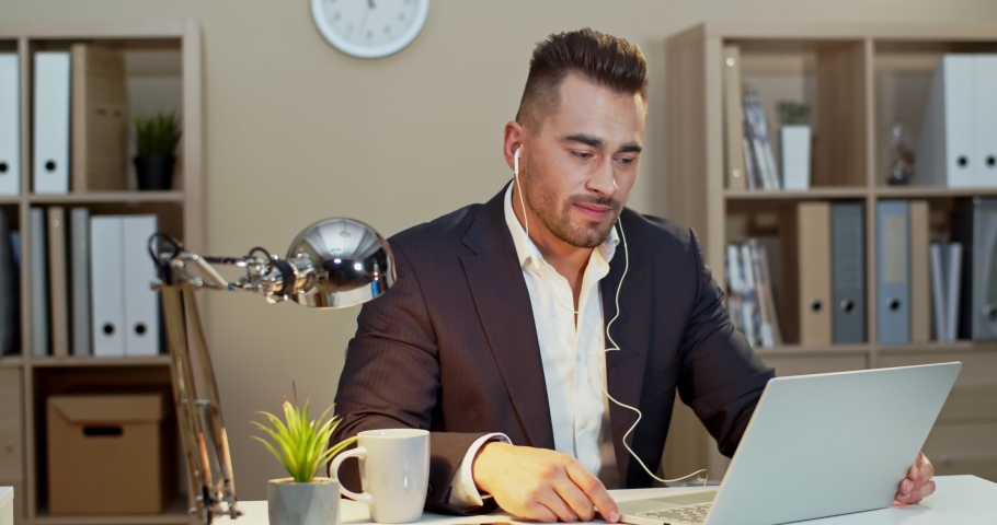 Good looking and happy Caucasian man in business style and headphones having videochat on the laptop computer in his ofice via web cam. Indoors.