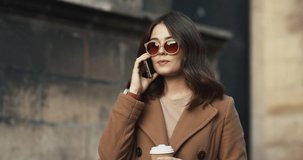 Close up of the young beautiful Caucasian woman in sunglasses and coat speaking on the mobile phone while walking outside with coffee to-go in hands.