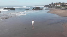 Aerial Top Down View of of happy woman in white dress running on coastline and making video of herself with drone on beach with black sand. Vacation in Bali.