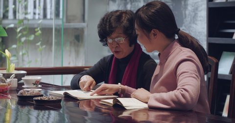 Chinese senior profesor teaching young grace woman traditional literature reading book together, senior mentor instructing student Chinese culture discussing talking together at home or library