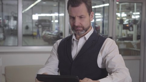 Portrait of young Caucasian male manager of repair shop sitting with tablet at the background of cars. Successful man waiting for car after inspection. Warranty service, jobbing shop.