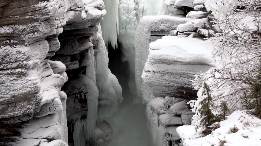 Aerial view of a nearly frozen waterfall in panning right with bare shrub right, Jasper National Park, Alberta Canada