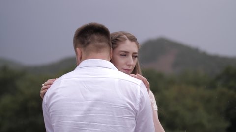 pretty bride hugs muscular fiance in white shirt standing against blurry hills with forests close view slow motion