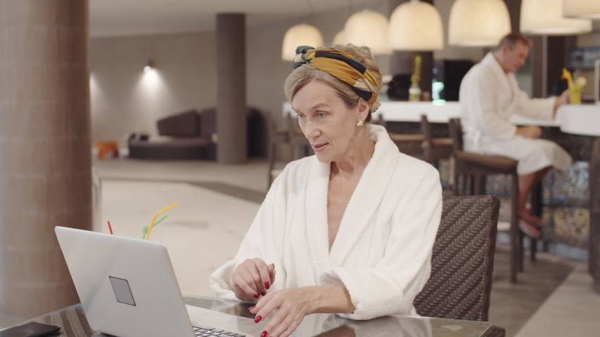 Pan shot of senior attractive woman wearing white bathrobe sitting at table in swimming center, using laptop and drinking cocktail