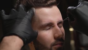 Close up of barber's hands in black gloves doing threading procedure and correcting shape of eyebrows to handsome bearded client sitting in chair. Concept of styling and care