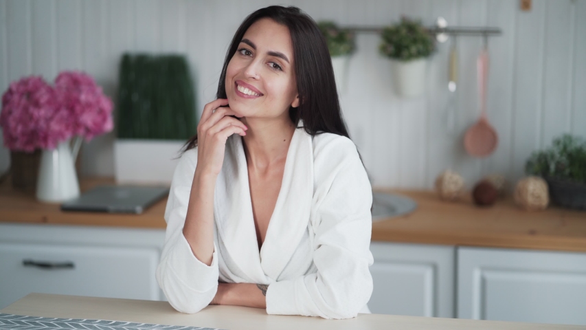 Portrait of cute young woman with beautiful smile sits in kitchen at home, looks at camera. Female nature beauty