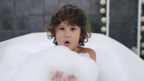 Portrait face of small cute boy with dark curly hair which playing fun in the with water and bath foam during bathing in the bathub. Slow motion
