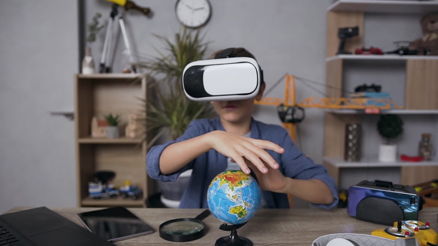 Slow motion of handsome modern smart teenage boy studing the structure of earth using augmented reality headset and little mockup of globe