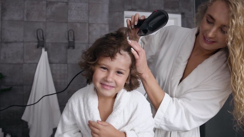 Caring young mom with long curly hair dries curly hair with a hairdryer her little happy son after taking shower at bathroom. Mom with son, happy family. Slow motion