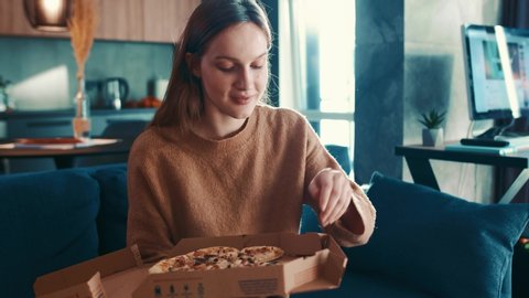 Cheereful european young woman in brown sweater sitting on sofa at modern living room and greedily eating tasty pizza, enjoying and smiling. Fast food addiction. Slow motion.