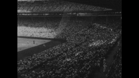 1940s: Baseball stadium. Spectators sit and stand. Players run onto field. Pitcher throws ball. Player hits ball and runs bases.