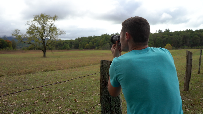 Young man taking a picture of a tree growning in a fenced meadow with a dramatic overcast sky in the background