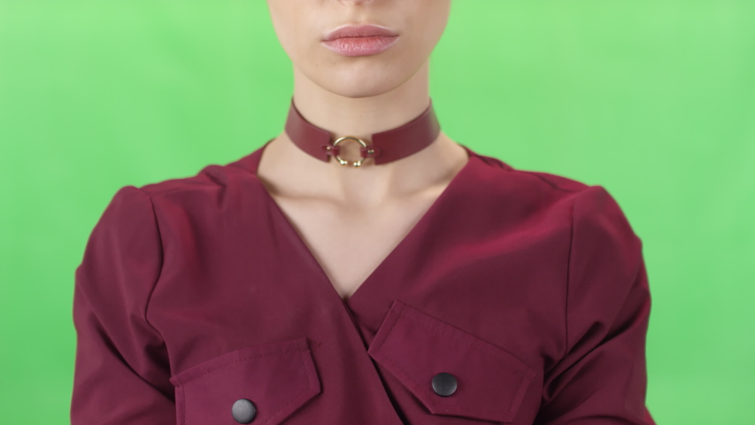 Close up midsection shot of seductive young woman in marsala outfit and choker holding hand on her chin and posing for camera against green chroma key background