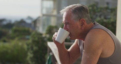 Side view close up of a senior Caucasian man relaxing at home , standing on his balcony in the sun, drinking a cup of coffee in the morning,Social distancing and self isolation in quarantine