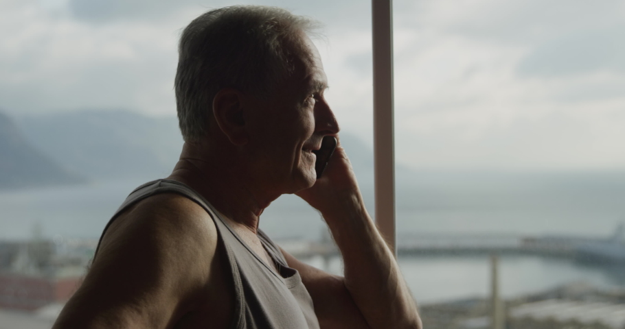 Side view close up of a senior Caucasian man relaxing at home wearing a vest, standing by the window talking on a mobile phone and laughing, slow motion