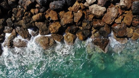 Panning aerial shot of seawall, waves break on rough stones. Beautiful turquoise color of crystal clear sea water. Sun light reflection flash in small puddles on wet rocks