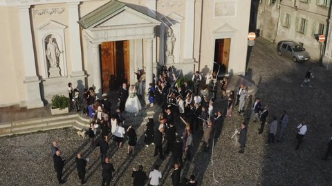 Top aerial view of newly wedded couple standing by ancient cathedral on wedding ceremony. Drone flying over bride groom and guests scattering confetti on celebration. Old Europe town destination