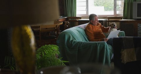 Side view of a senior Caucasian man relaxing at home in his living room, sitting on the sofa using a laptop computer and stroking his pet dog, slow motion