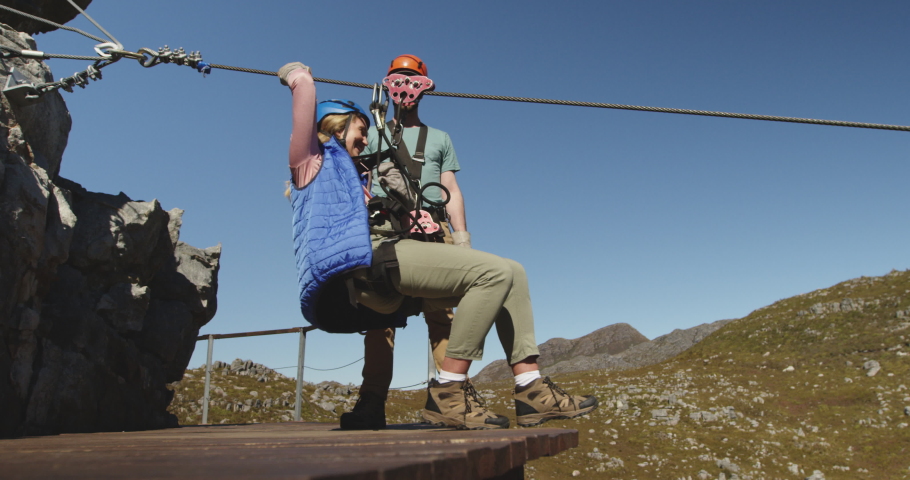 Side view of a young Caucasian woman zip lining on a sunny day in mountains while a young Caucasian man is helping her and then watching her ride, slow motion. Adventure Vacation in South Africa