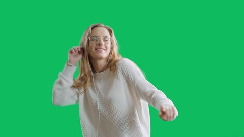Happy young woman in earphones is listening to music with smart phone, dancing and smiling in a green screen background. The positive teenager girl with glasses and headphones in chroma key background