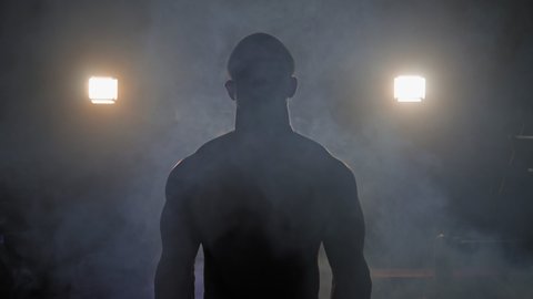 Muscular man doing crossfit training in a dark shadowy gym lifting weights holding a barbell at waist level in a health and fitness concept, movement round shot