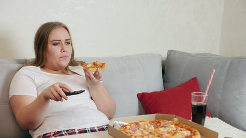 An overweight girl with a big appetite eats pizza while sitting on the sofa with TV remote control in hand. On the table large box of pizza and glass with cola