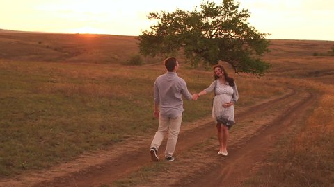 A happy pregnant woman and her husband are walking along a country road in a field at sunset in summer. Slow motion.