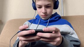 Teenager boy plays video games on the mobile phone. Young man cyber sportsman sitting on the armchair with  headphones and smartphone