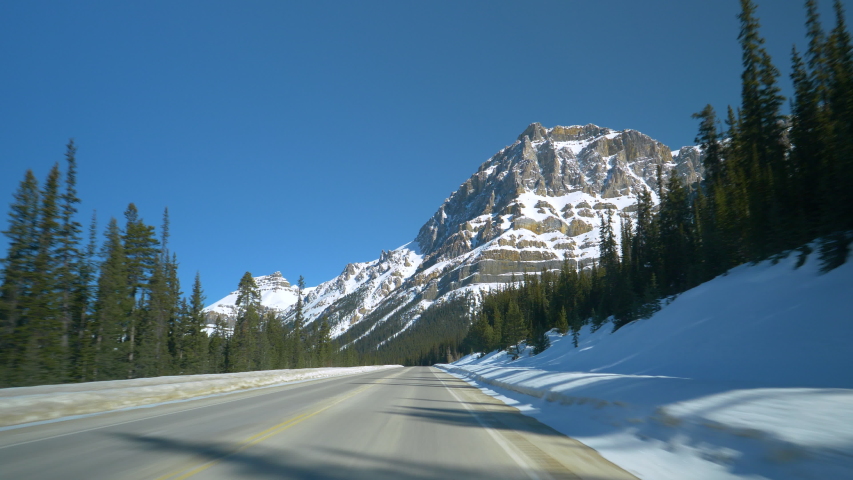 POV: Driving along an empty asphalt road leading through the beautiful Banff National Park. Scenic first person view of driving down the Icefields Parkway on a sunny winter day. Road trip scenery.