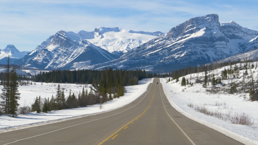 SLOW MOTION: Scenic view of the snowy Canadian Rockies as car disappears in the distance. Driving along the famous Icefields Parkway tourist route on sunny winter day. Picturesque Banff National Park