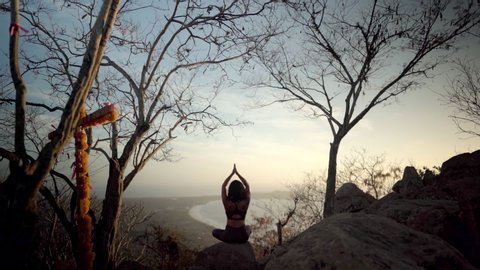 Young woman sits in namaste pose meditating on a beautiful mountain as sun sets in the distance