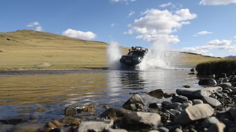 4WD off-road vehicle is crossing the water with white splashes with the hill and blue sky on background. Altay landscape with mountain river on a cloudy day