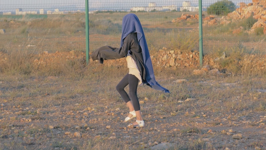 homeless child walks near protected area. skinny refugee girl head covered quilt goes along the iron fence state border. concept: war aftermath and political refugees.
