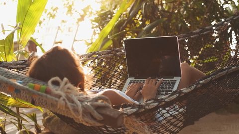 Beautiful young european woman seated in a hammock working with a laptop on a sunset background. Concept remote worker, position freelance entrepreneur.