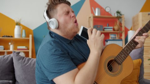 Slow motion of happy young man playing the guitar singing in smartphone wearing wireless headphones at home. Modern technology, people and fun concept.
