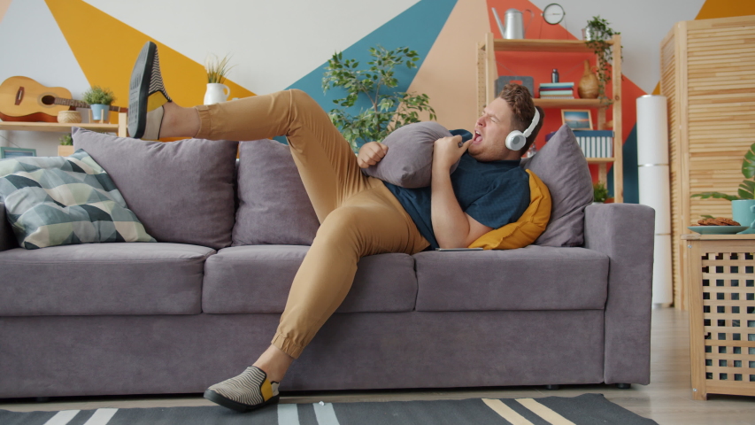 Funny young man in headphones is enjoying music singing playing the guitar on pillow having fun at home on sofa. Modern technology and lifestyle concept.