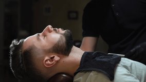 Close up of male barber in black gloves shaving client with sharp steel razor. Handsome guy lying on couch with closed eyes getting new trendy beard in modern barbershop