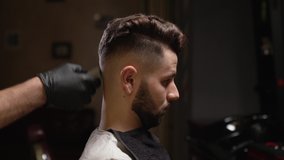 Side view of male hipster getting haircut by barber at modern salon. Professional hairdresser in black gloves cutting hair of client with trimmer machine.
